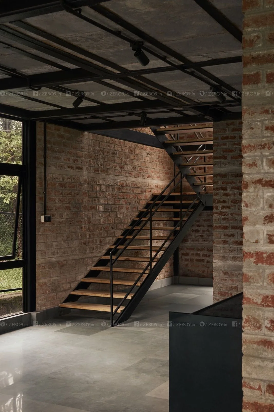 Interior staircase — open-tread timber and steel beside exposed brickwork, with glazed windows framing the forest